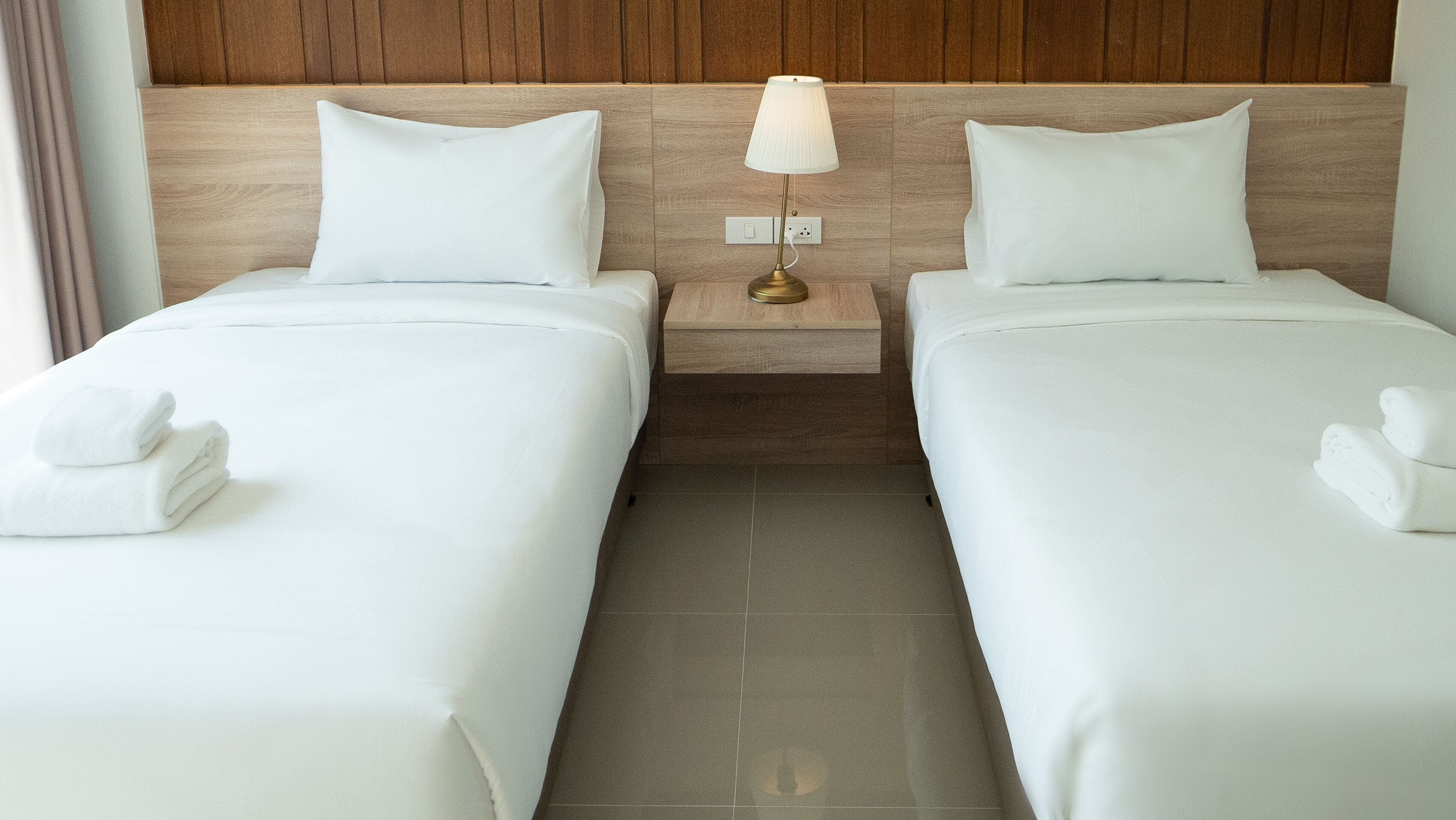 A hotel room with two single beds, a nightstand with a lamp, and a tiled floor.