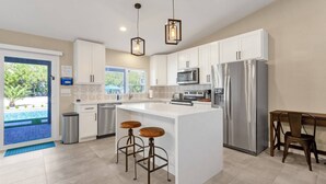 A modern kitchen with white cabinets, stainless steel appliances, a central island with stools, and a view of a swimming pool through a glass door. - Heated Pool w/ swim up bar seating & Spa, Pool Table, Basketball Court, Putting green + FREE Golf! (Scottsdale)