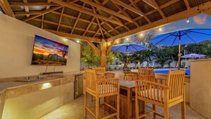 An outdoor patio with wooden chairs and a table, a bar area with a sink and a refrigerator, and a pool in the background. - Heated Pool w/ swim up bar seating & Spa, Pool Table, Basketball Court, Putting green + FREE Golf! (Scottsdale)