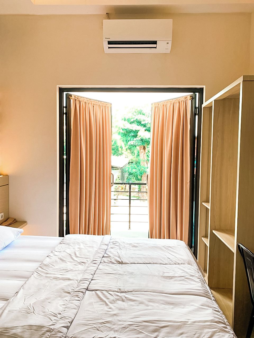 A hotel room with a bed, an air conditioning unit, a wardrobe, and a view of the outdoors through a doorway.