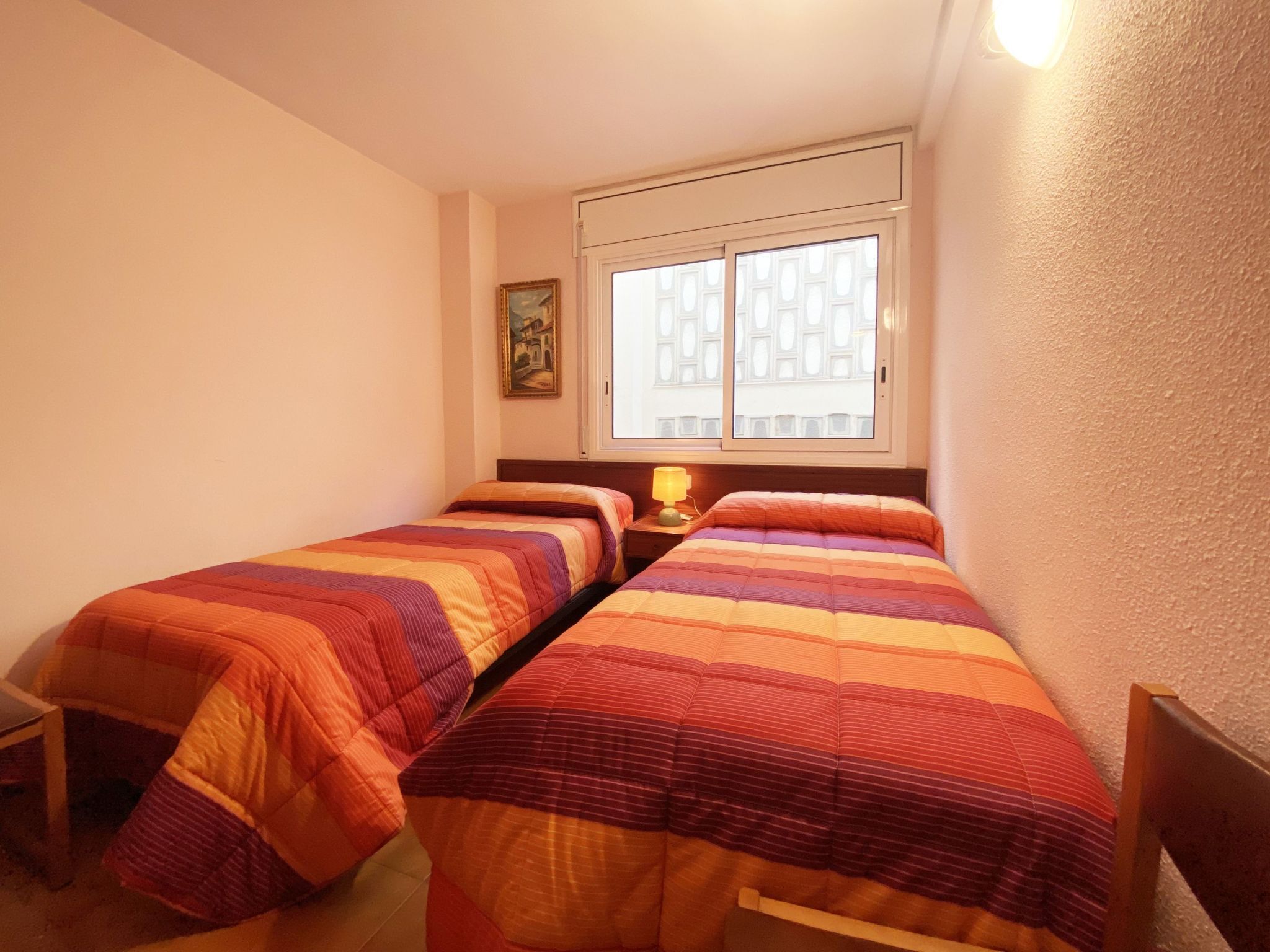 Une chambre avec deux lits, une petite table avec une lampe, un tableau au mur et une fenêtre donnant sur des immeubles.