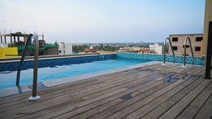 Una terraza de madera con piscina, escaleras metálicas y vista a edificios al fondo.
