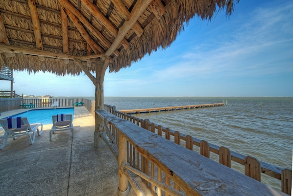 Un espace piscine situé au bord de la plage, doté d’une terrasse en bois, de chaises longues et d’une structure à toit de chaume.