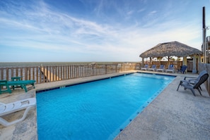 Une piscine avec une terrasse en bois, des chaises longues et un parasol en chaume.