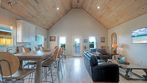A spacious loft-style living area with a high ceiling, a dining table with chairs, a sofa, and a television mounted on the wall. - Bay Haven w/ Shared Boat Dock & Waterfront Pool | Big Trout by AvantStay (Corpus Christi)