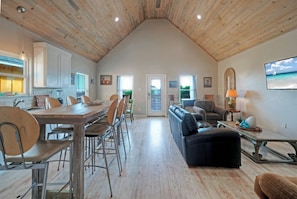 A spacious loft-style living area with a high ceiling, a dining table with chairs, a sofa, and a television mounted on the wall.