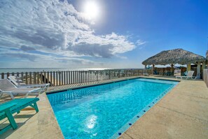 Une piscine dotée d’une terrasse en bois et de chaises longues, une structure à toit de chaume et une clôture en bois offrant une vue sur l’océan.
