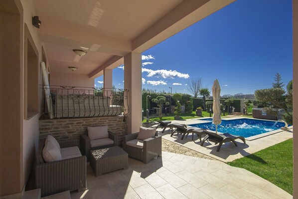 A covered outdoor area with wicker furniture, a pool, and a view of a landscaped garden.
