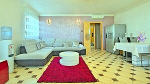 Una sala de estar con un sofá modular, un tapete rojo, un dispensador de agua, un refrigerador y una mesa de comedor con mantel blanco.