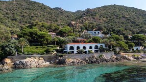 A coastal property with a white building featuring arched windows, surrounded by lush greenery and rocky terrain. - VILLA DOLCE VITA- 8 bedrooms, sea view, swimming pool, (Ajaccio)
