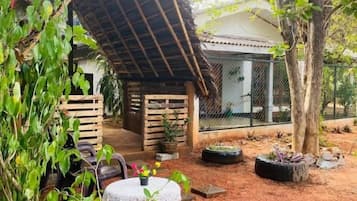A covered outdoor seating area with a table and chairs, surrounded by plants and trees.