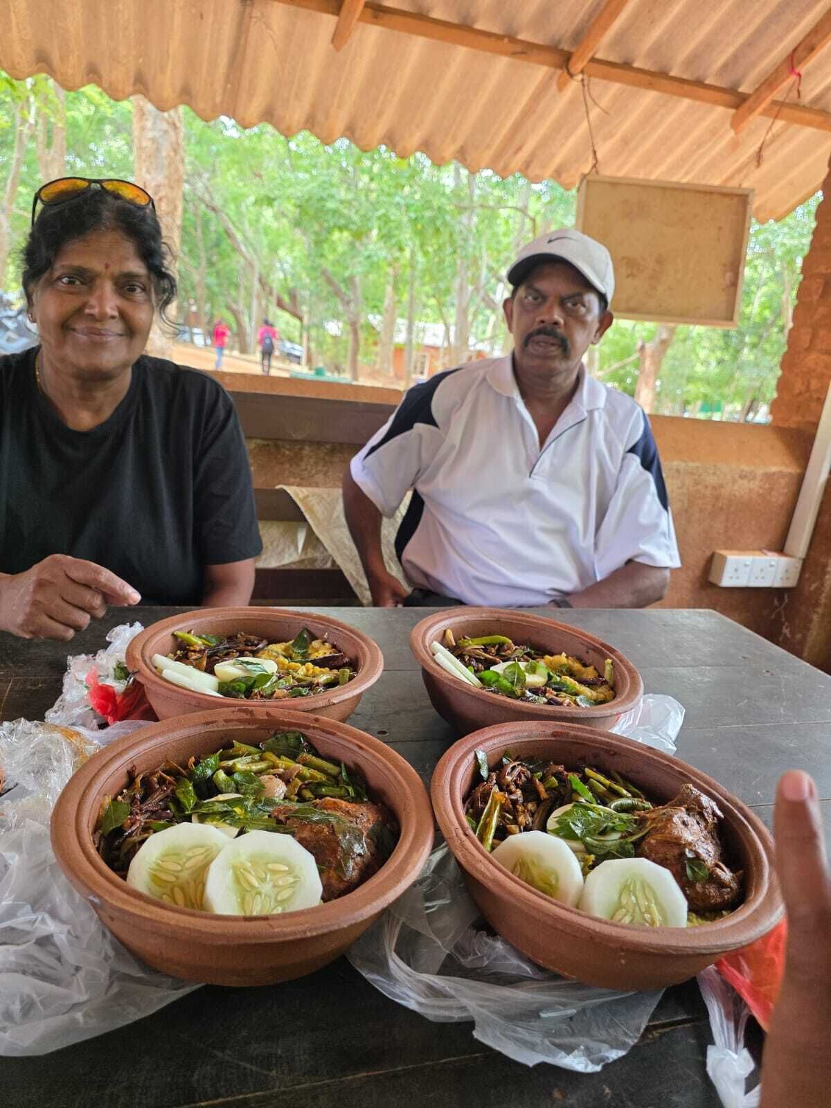 Two people seated at a table with four terracotta dishes containing food, under a wooden structure with a view of trees.