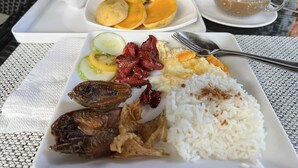 Uma refeição com arroz, peixe frito, carne fatiada, ovos e uma salada de frutas em um prato branco, com uma xícara de café em uma bandeja ao fundo.