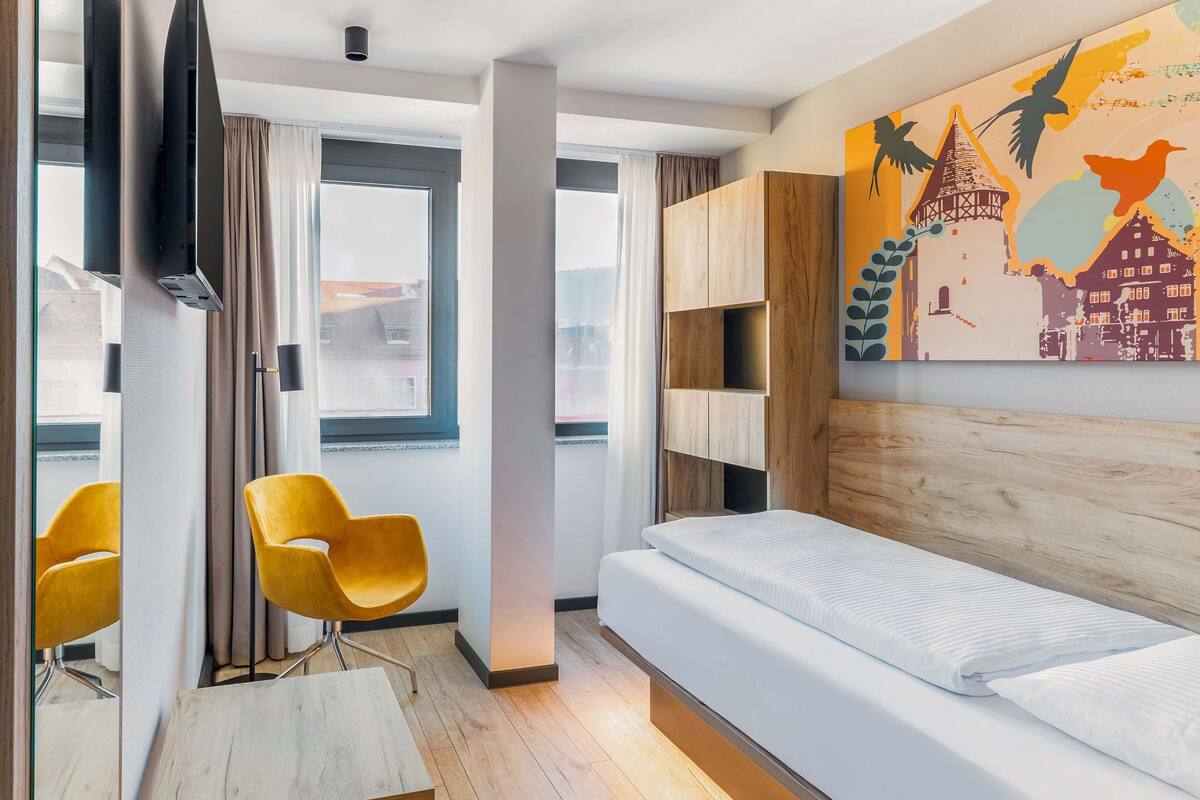 Una habitación de hotel moderna con una cama, cabecera de madera, televisor montado en la pared, una silla amarilla y una pintura de paisaje urbano.