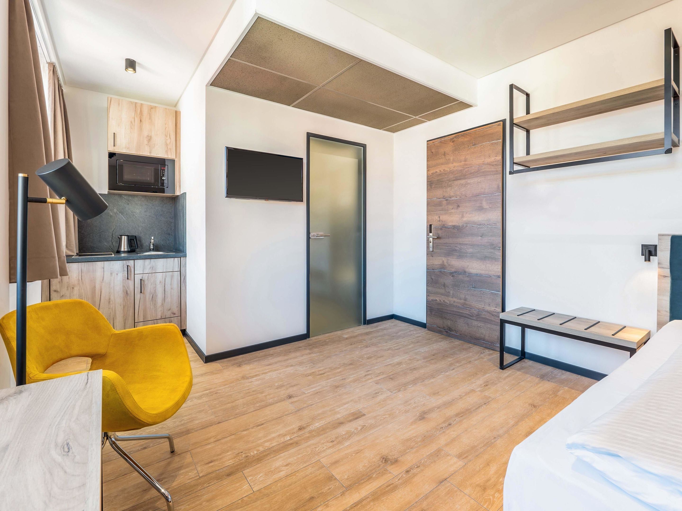 A compact hotel room with a kitchenette, a bed, a yellow chair, and a built-in bench.