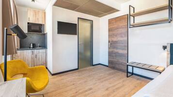A compact hotel room with a kitchenette, a bed, a yellow chair, and a built-in bench.