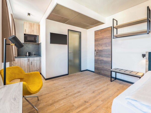 Une chambre d’hôtel compacte comprenant un coin cuisine, un lit, une chaise jaune et une banquette intégrée.