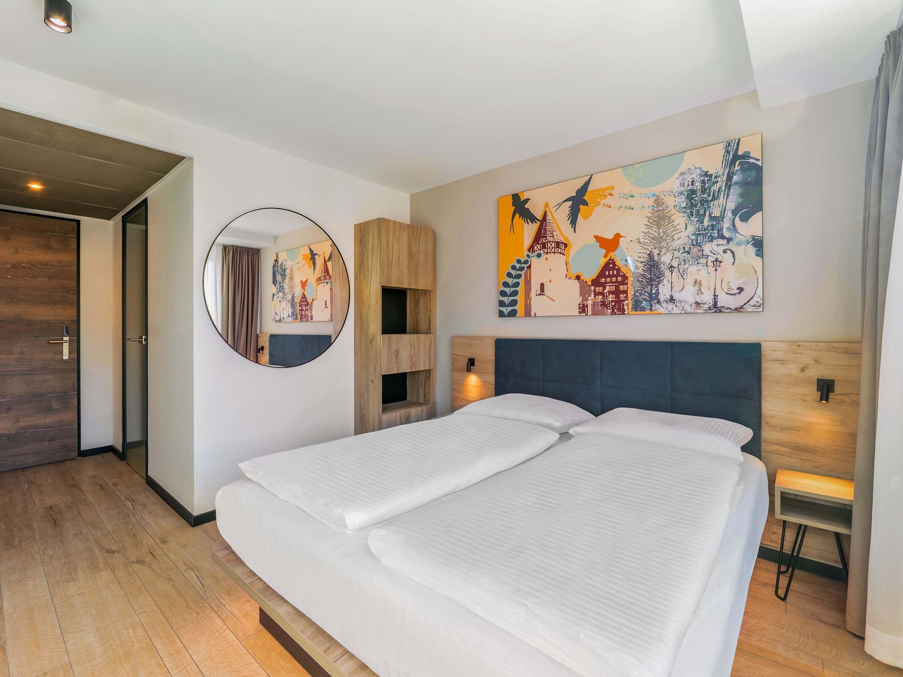 A modern hotel room with a large bed, a round mirror, and a colorful cityscape painting on the wall.