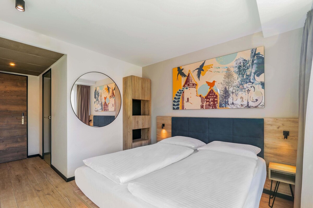 Une chambre d’hôtel moderne avec un grand lit, un miroir rond et un tableau coloré représentant un paysage urbain accroché au mur.