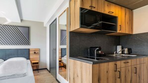 Una habitación de hotel moderna con kitchenette de madera, cama con ropa de cama blanca, una pequeña zona de estar y un ventanal grande.