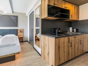 Une chambre d’hôtel moderne avec une kitchenette en bois, un lit avec une literie blanche, un petit coin salon et une grande fenêtre.