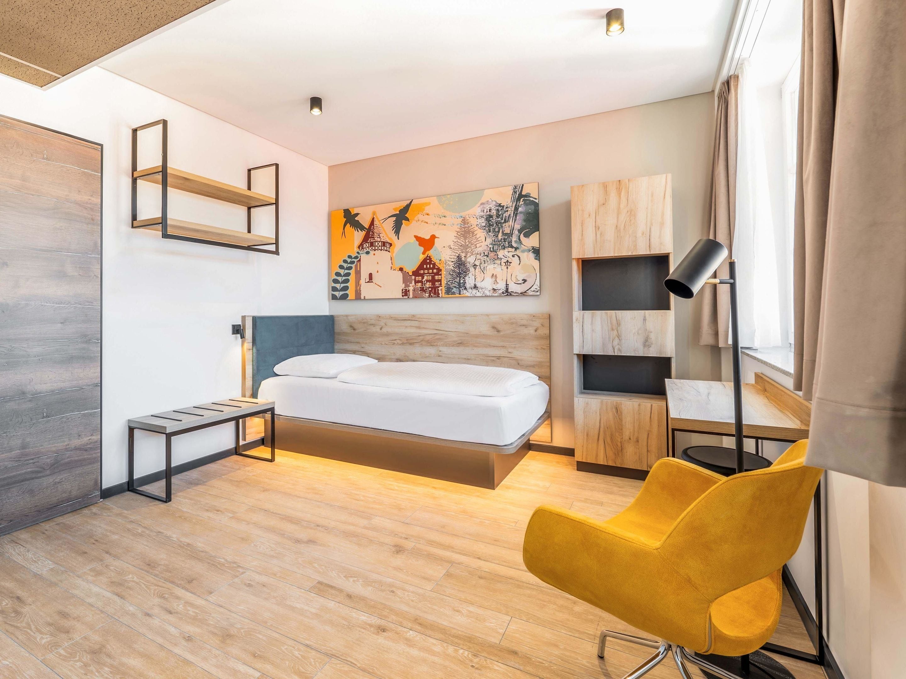 A modern hotel room with a wooden floor, a bed with a wooden headboard, a yellow chair, a desk with a lamp, and a painting on the wall.