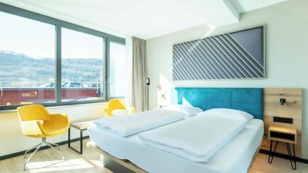 Una camera d'hotel con una grande finestra, un letto con lenzuola bianche, una testiera blu, una sedia gialla e un tavolino.