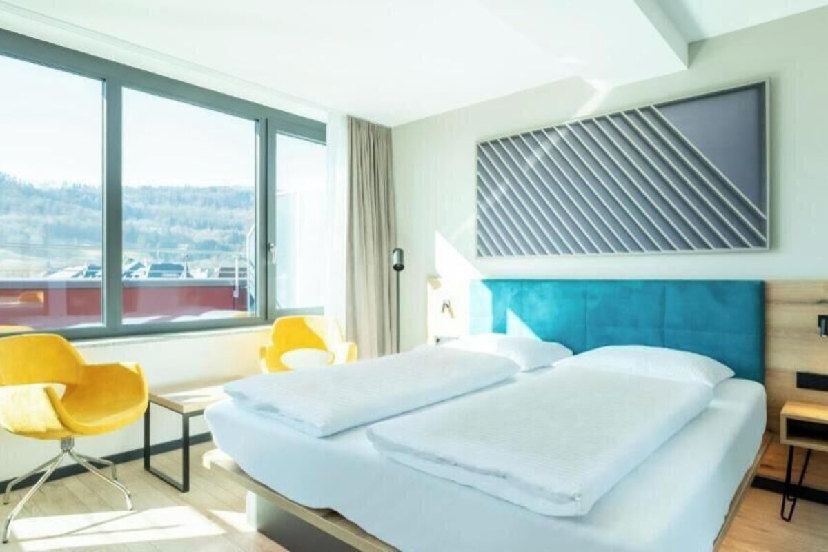 Une chambre d’hôtel avec une grande fenêtre, un lit avec du linge de lit blanc, une tête de lit bleue et une chaise jaune.