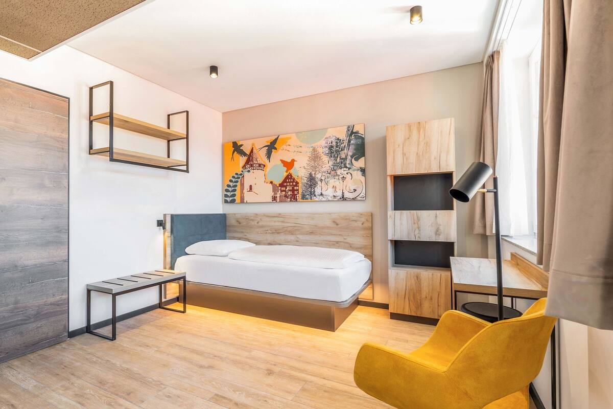 Una habitación de hotel moderna con piso de madera, una cama con cabecera de madera, una silla amarilla, un escritorio con lámpara y un cuadro en la pared.