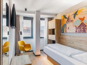 A modern hotel room with a bed, a wooden headboard, a TV mounted on the wall, a yellow chair, and a cityscape painting. - B&B HOTEL Albstadt-Hbf (Albstadt)