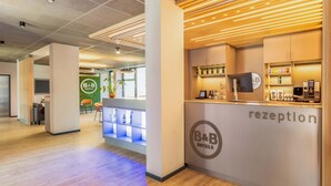 Area reception di un hotel dal design moderno, con scrivania, parete verde con il logo dell'hotel e zona relax con una pianta.