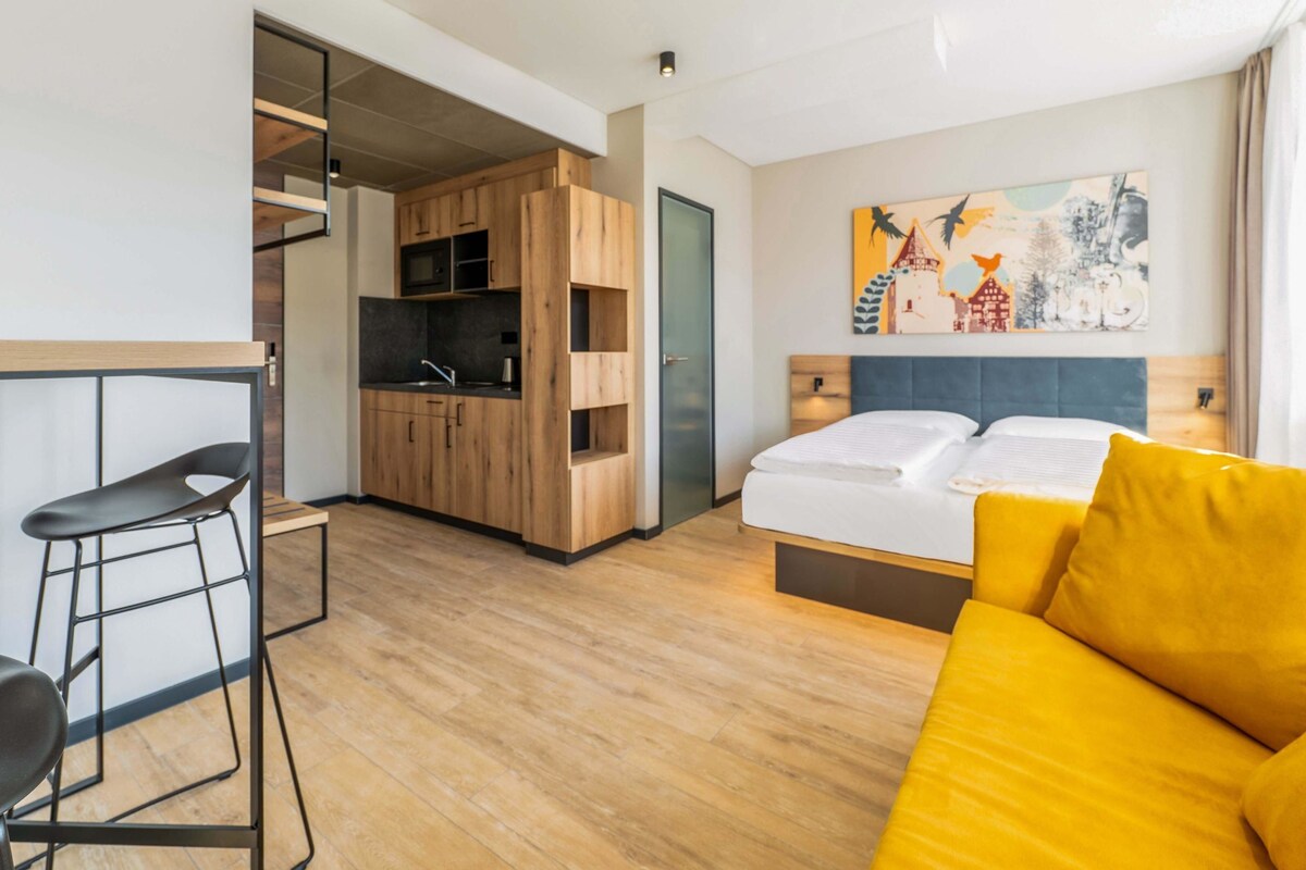 Une chambre d’hôtel compacte avec une kitchenette, un lit avec des oreillers jaunes, une chaise moderne et un tableau au mur.