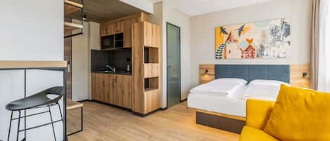 A compact hotel room with a kitchenette, a bed with yellow pillows, a modern chair, and a painting on the wall.