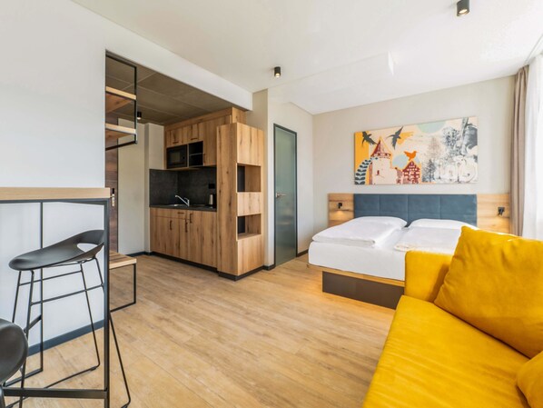 A compact hotel room with a kitchenette, a bed with yellow pillows, a modern chair, and a painting on the wall.