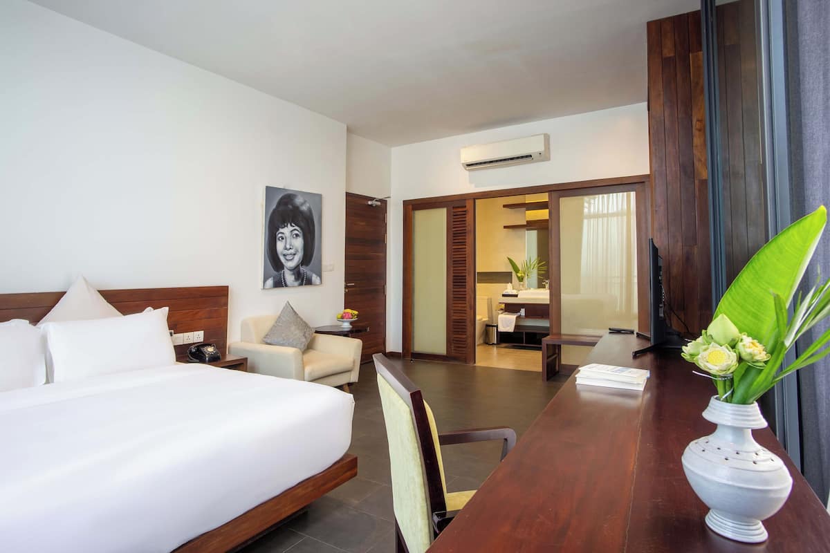 A hotel room with a large bed, a desk with a vase of flowers, a chair, a sofa, and a bathroom visible through an open door.