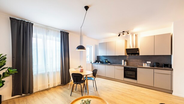 A modern kitchen with a dining area, wooden flooring, and a large window with curtains. - Glück Auf Appartements Herkulesstraße (Essen)