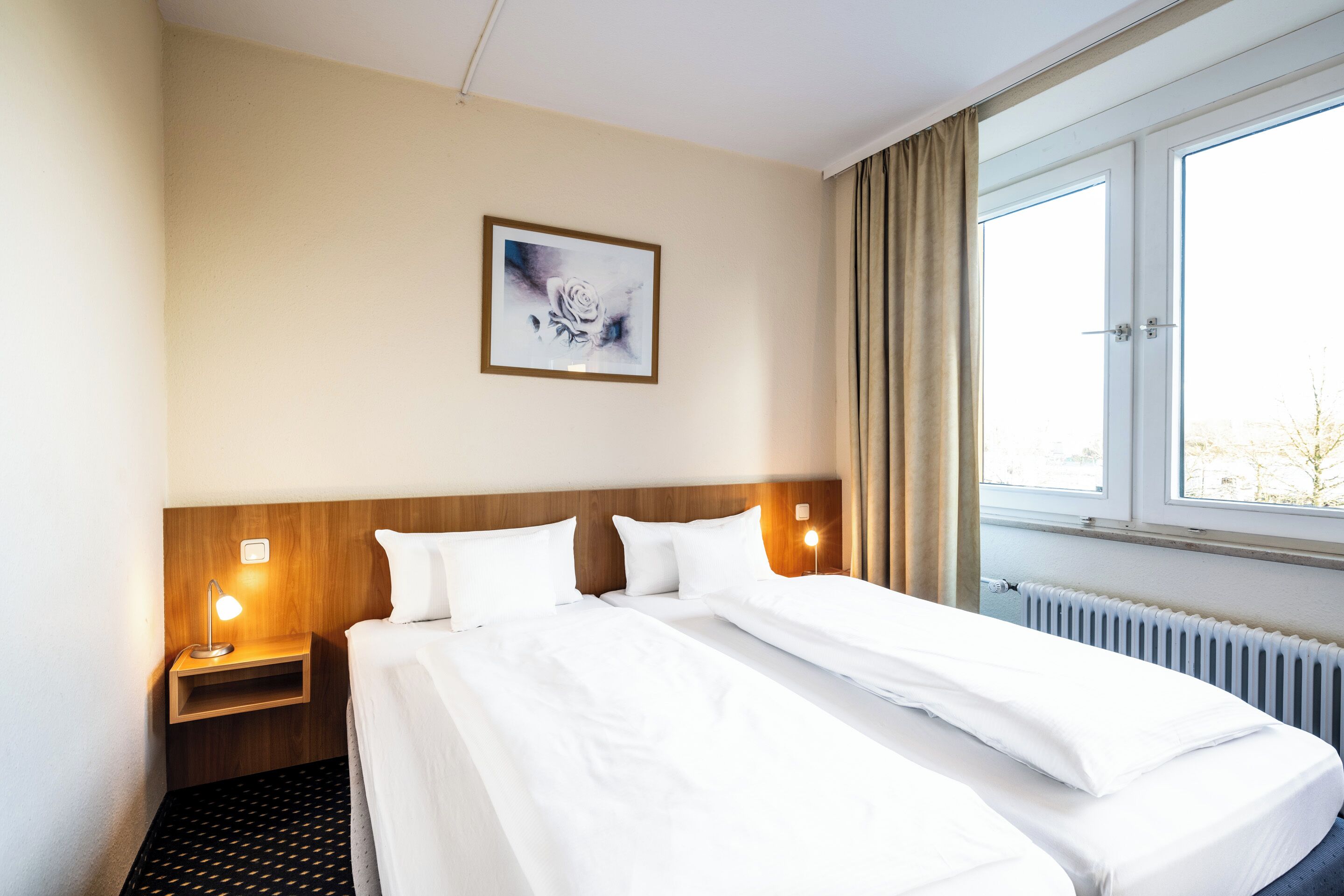 Ein Hotelzimmer mit einem großen Bett, einem Nachttisch mit Lampe, einem gerahmten Bild an der Wand und einem Fenster mit Vorhängen.