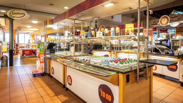 Uma cafeteria com um balcão de vidro repleto de bebidas engarrafadas, uma máquina de café e um aviso que diz "TO GO".