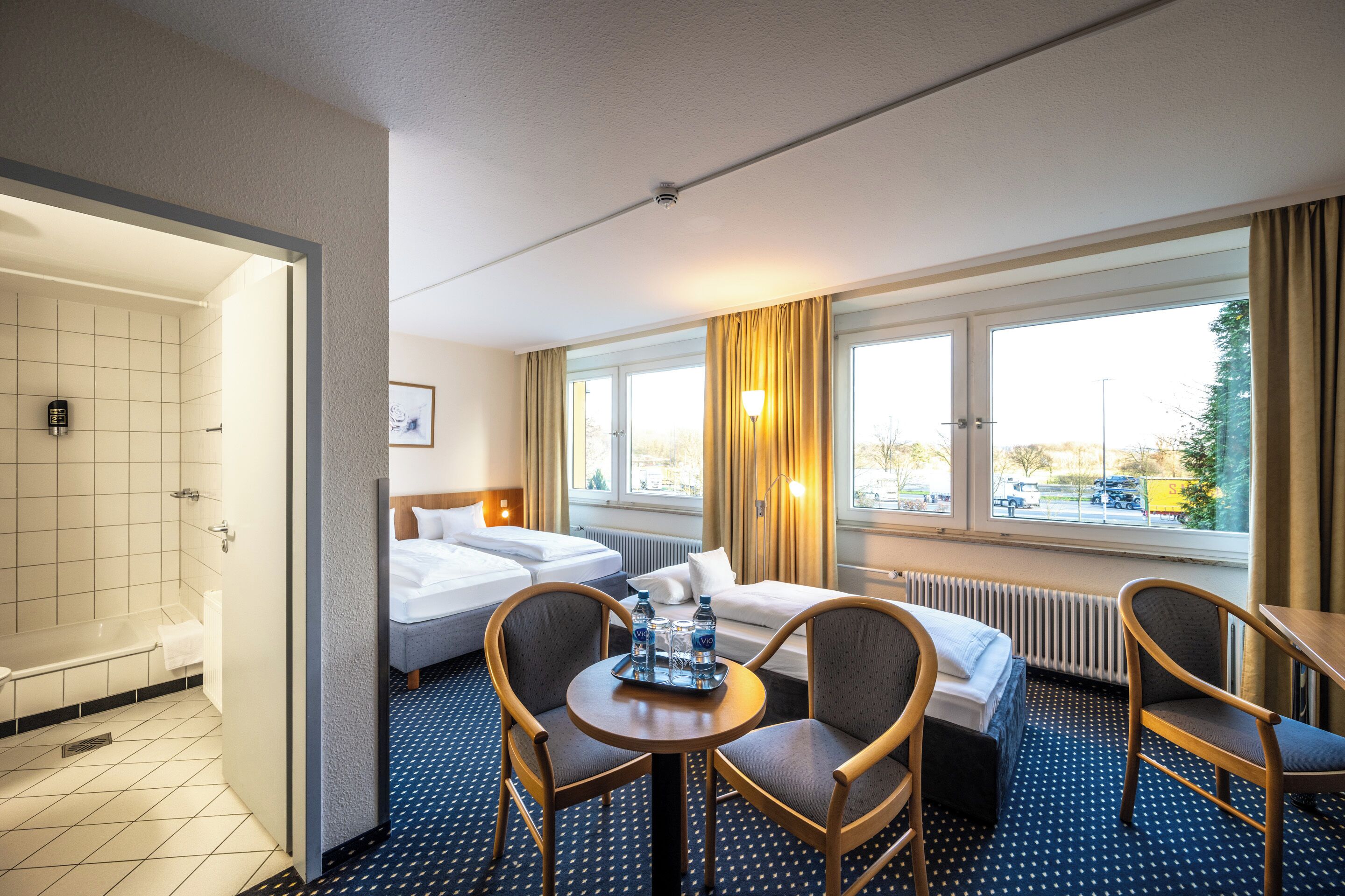 A hotel room with a bed, two chairs, a table with water bottles, a window with a view, and a bathroom with a bathtub and toilet.