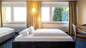 Quarto de hotel com duas camas, uma escrivaninha e vista para árvores através de amplas janelas.