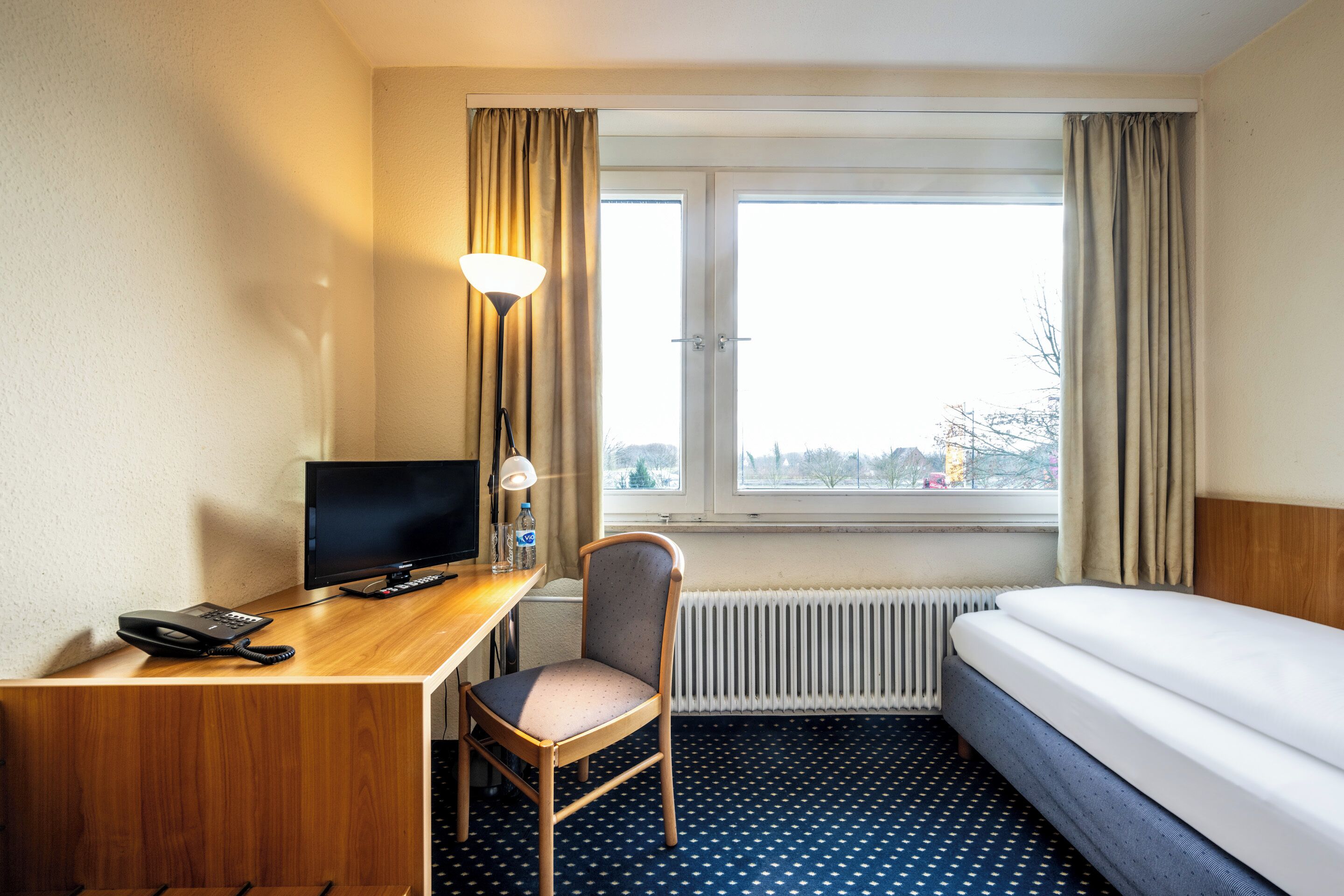 Une chambre d’hôtel avec un bureau en bois, une chaise, un lit, une télévision et une fenêtre avec des rideaux.