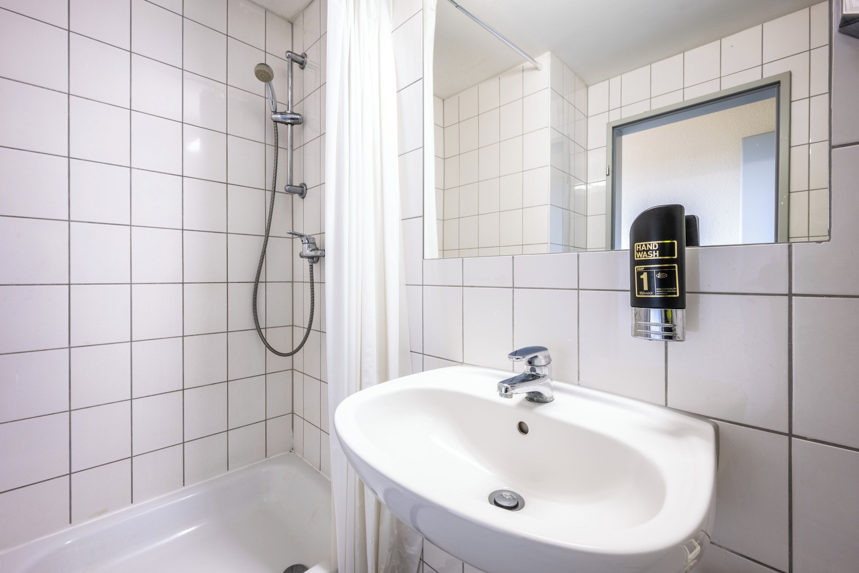 Une salle de bain avec des carreaux blancs, un lavabo blanc, une douchette à main et un distributeur de savon liquide.