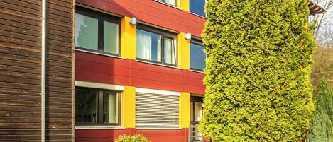 A multi-story building with a yellow facade, a red and yellow stripe, and a large green tree in front.