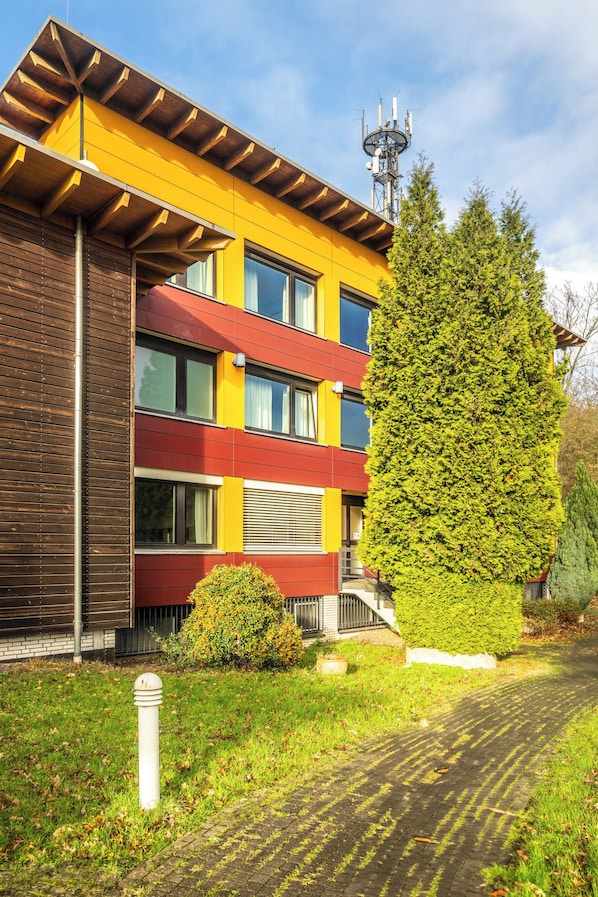Un edificio de varios pisos con fachada amarilla, una franja roja y amarilla, y un gran árbol verde en la parte delantera.