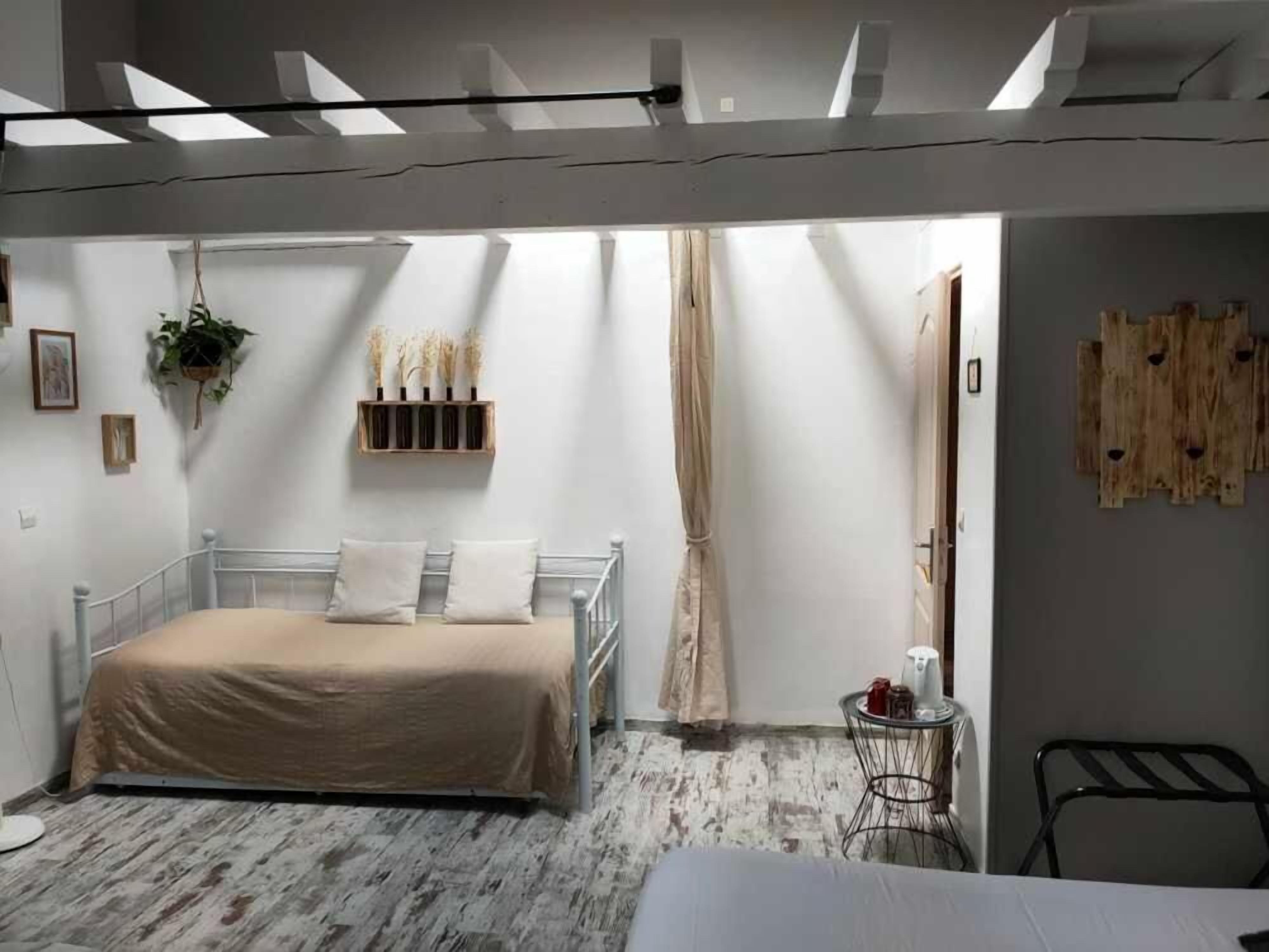 A bedroom with a bed, a small table with a kettle, a hanging plant, and a wooden shelf with decorative items.