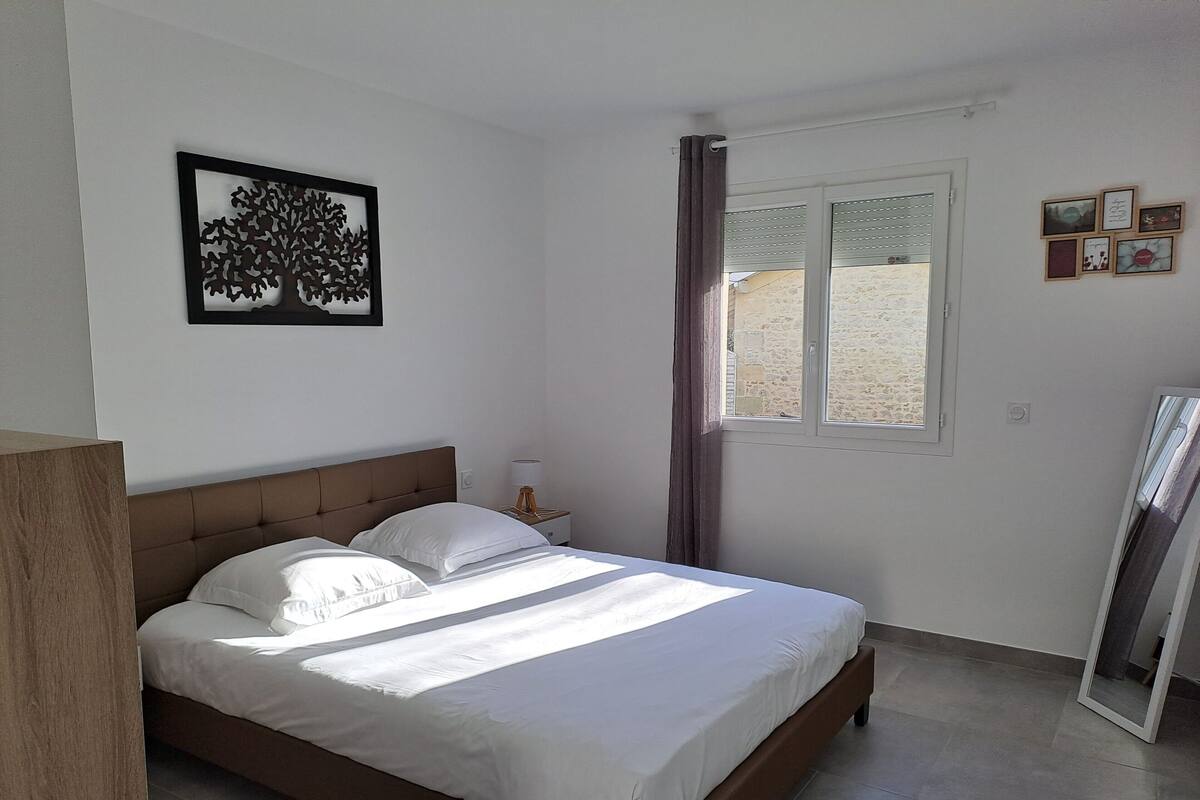 Un dormitorio con cama, mesita de noche, lámpara, ventana con cortinas e imagen colgada en la pared.