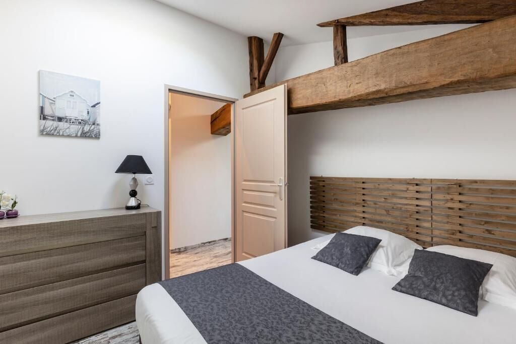 Un dormitorio con cabecera de madera, cama con ropa de cama gris y blanca, mesita de noche con lámpara y una puerta que da a otra habitación.
