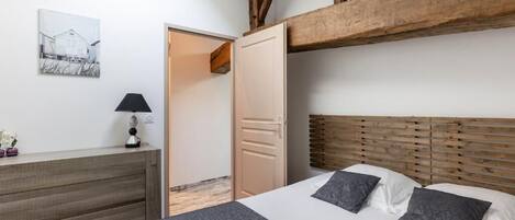 Un dormitorio con cabecera de madera, cama con ropa de cama gris y blanca, mesita de noche con lámpara y una puerta que da a otra habitación.