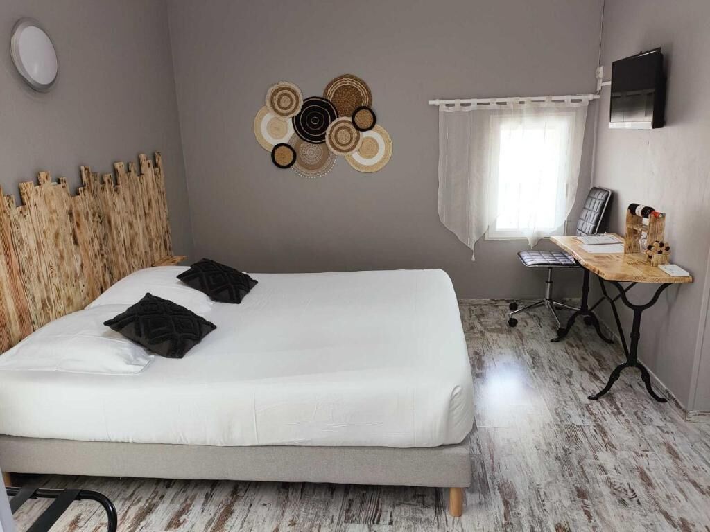 Une chambre à coucher avec un lit, une tête de lit en bois, un téléviseur fixé au mur, une petite table avec une lampe et une fenêtre ornée de rideaux légers.