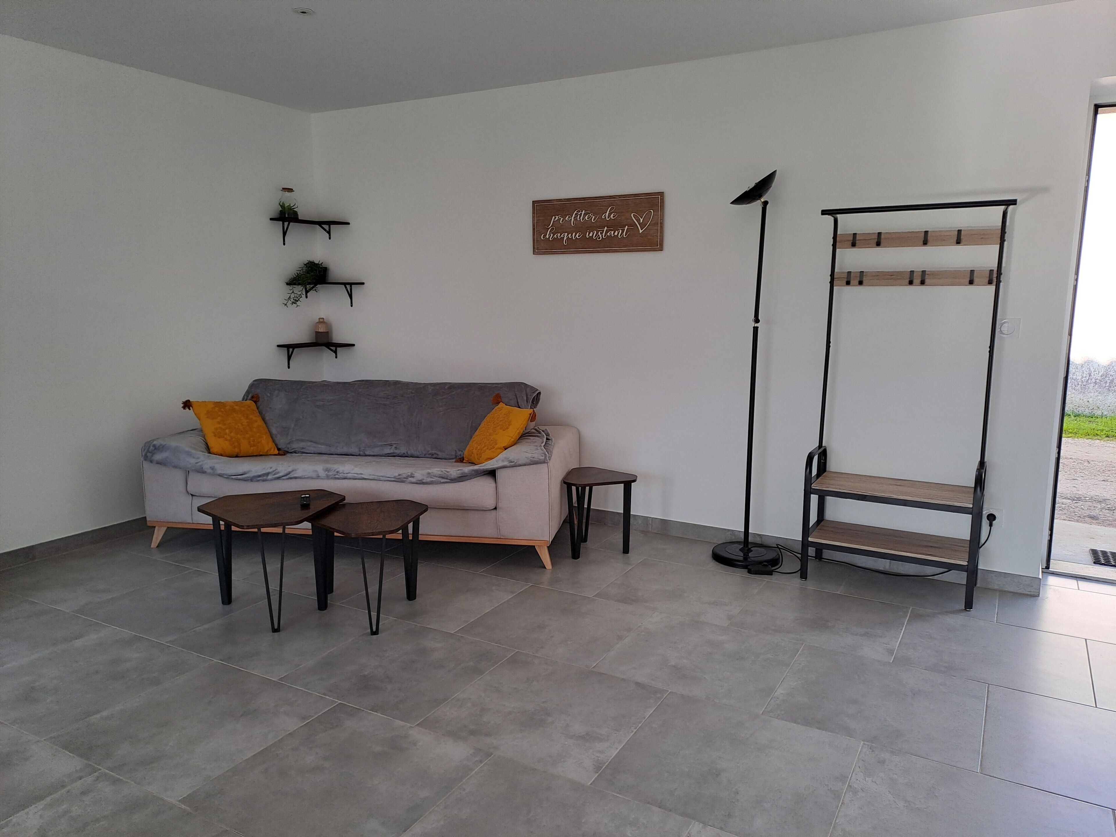 Ein modernes Wohnzimmer mit grauem Sofa, zwei kleinen Tischen, einer Stehlampe und einem Holzleitnerregal an einer weißen Wand.